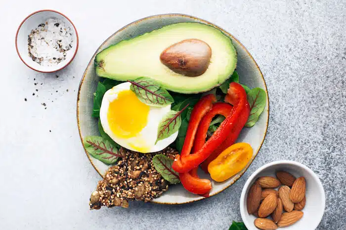 An overhead view of a plate with avocado, a fried egg, red pepper, grains and almonds